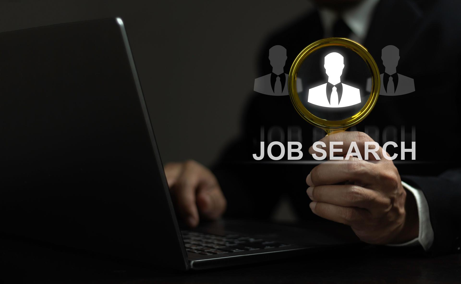 A man is holding a magnifying glass over a laptop screen that says Job Search